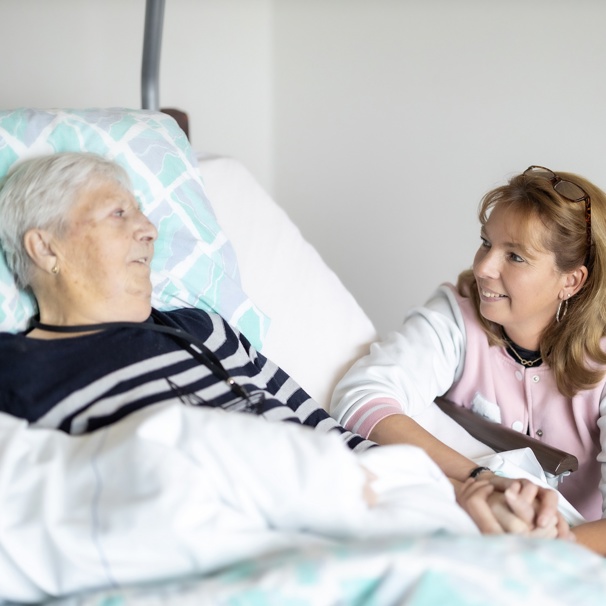 Een mevrouw ligt in bed. Een medewerker van het hospice zit bij haar, houdt haar hand vast. Ze kijken elkaar glimlachend aan. Afscheid nemen op een manier die bij je past kan kan in Hospice het Baken.