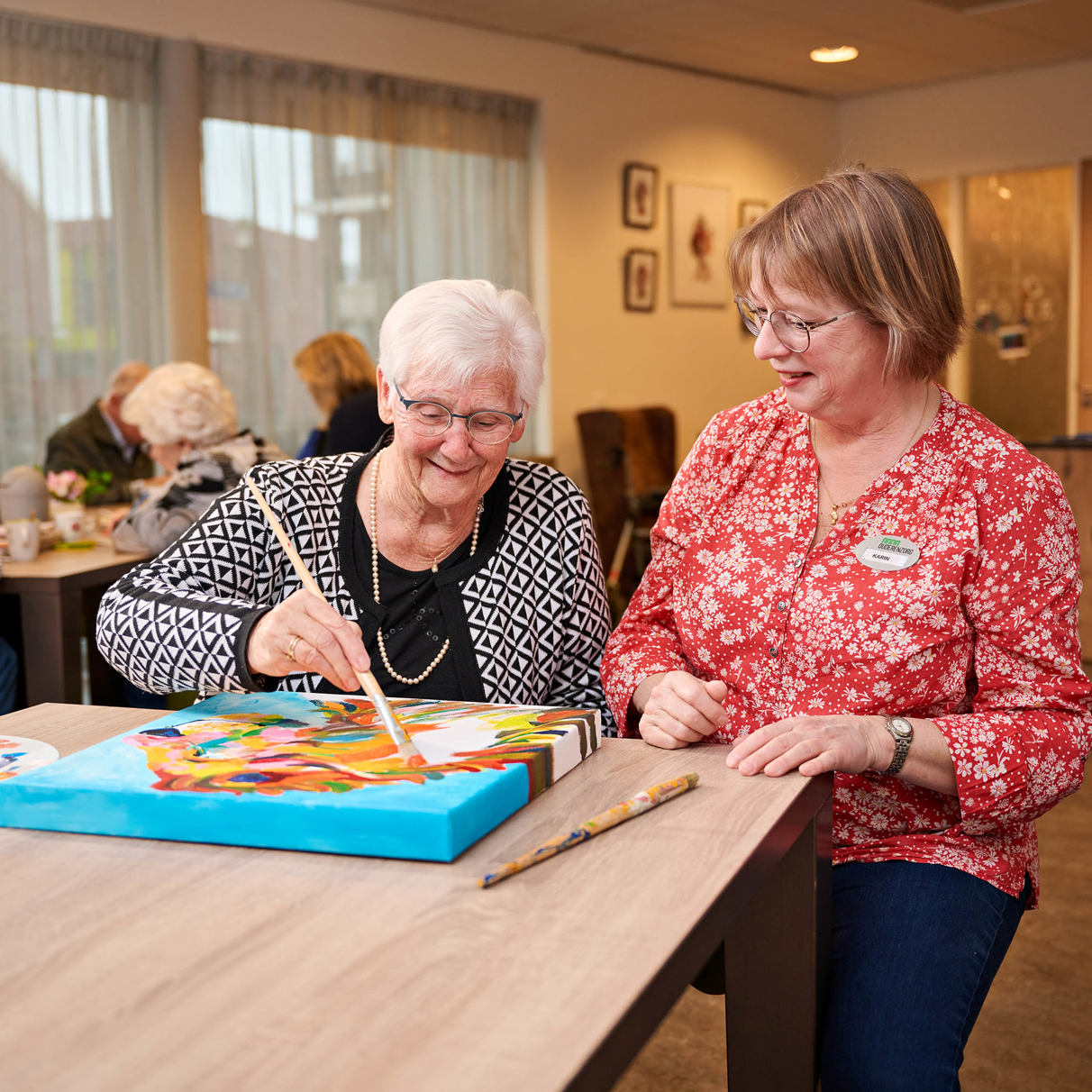 Twee dames zitten aan tafel, oudere dame schildert op schilderij dat op tafel ligt