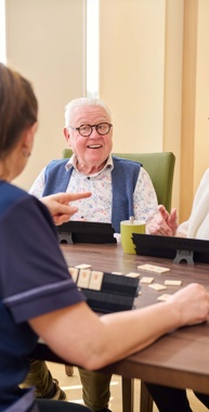Een oudere man en vrouw zitten samen met een medewerker van Anna Ouderenzorg aan tafel. Ze spelen een spelletje rummikub. Bij het Centrum voor Herstel en Revalidatie helpt je om na je ziekte of operatie weer sterker te worden zodat je weer goed voor jezelf kunt zorgen als je weer in je eigen huis bent.