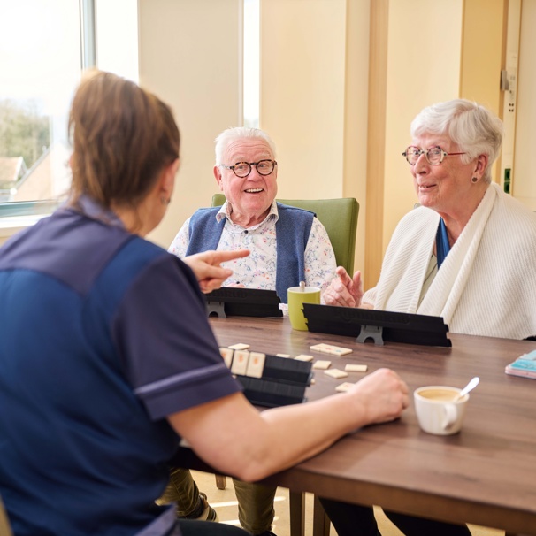 Een oudere man en vrouw zitten samen met een medewerker van Anna Ouderenzorg aan tafel. Ze spelen een spelletje rummikub. Bij het Centrum voor Herstel en Revalidatie helpt je om na je ziekte of operatie weer sterker te worden zodat je weer goed voor jezelf kunt zorgen als je weer in je eigen huis bent.