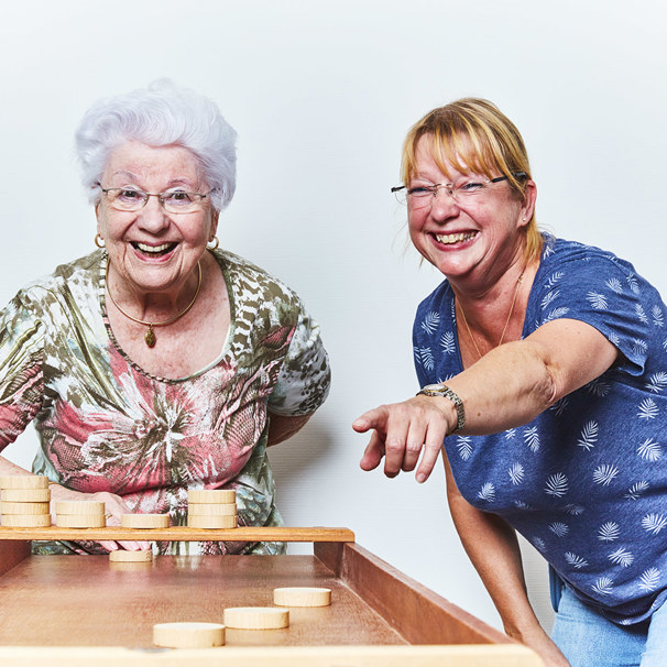 Activiteiten kalender Anna Ouderenzorg  Oudere dame staat achter een sjoelbak en andere dame staat ernaast, terwijl ze lachend naar de sjoelbak kijken