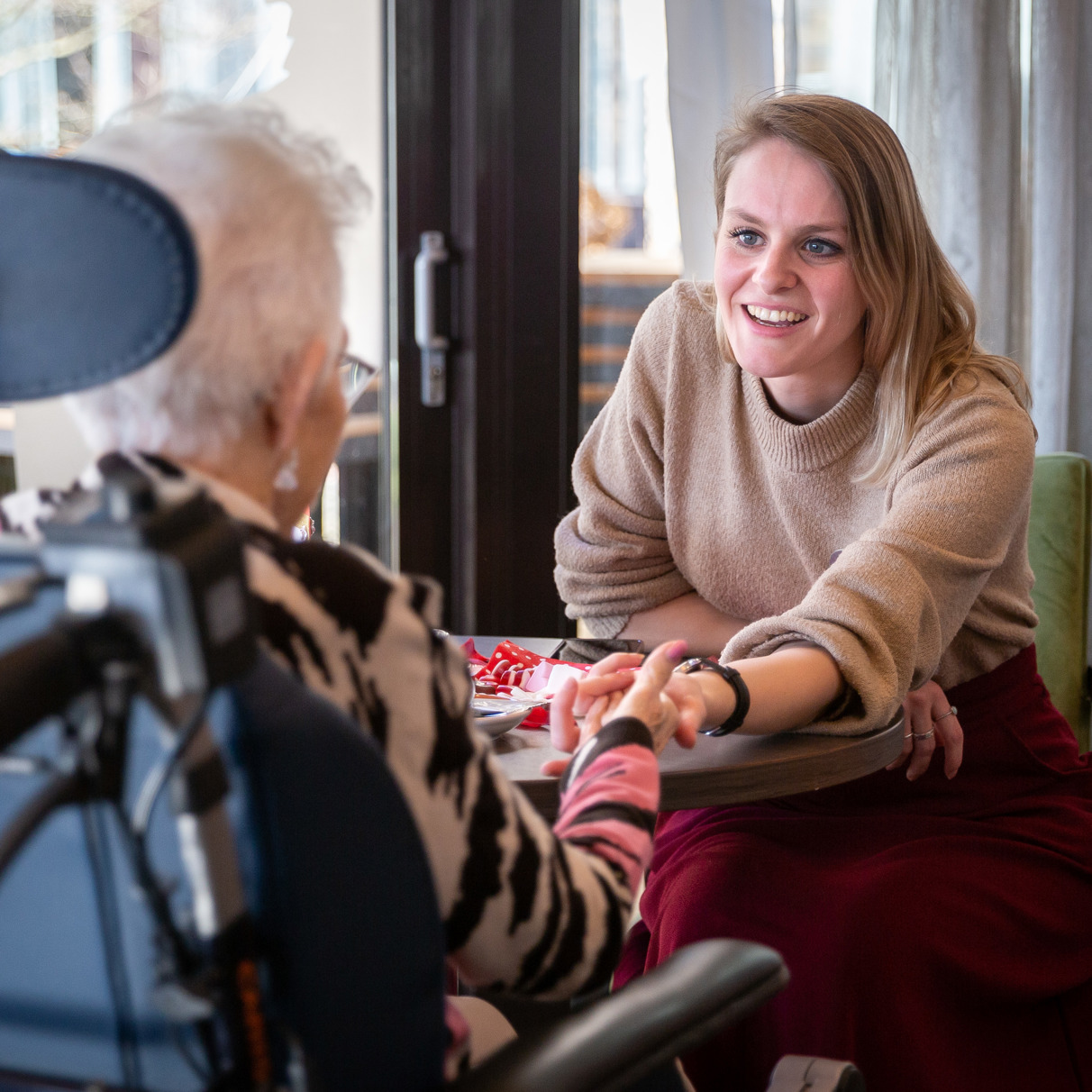 Activiteitenbegeleider zit voor mevrouw in rolstoel en houdt haar hand vast