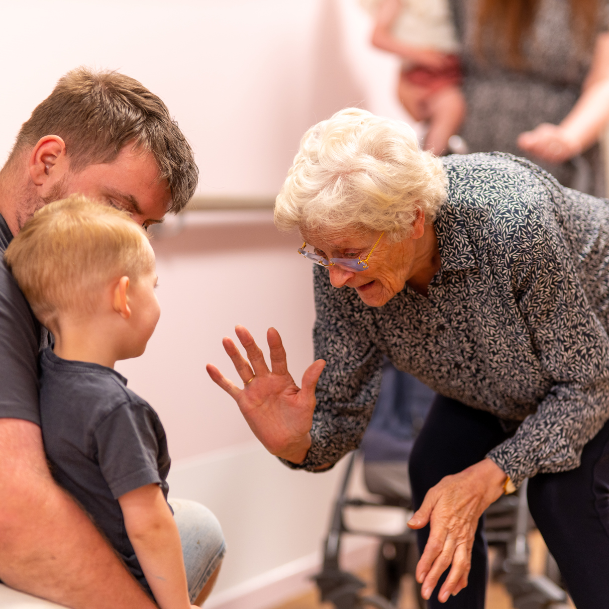 Oudere mevrouw bukt zich om een klein kind een high five te geven, die tegen zijn vader geleund staat