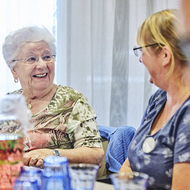 Activiteiten Anna Ouderenzorg kalender Twee dames lachend met elkaar in gesprek aan tafel, met daarop glazen met drinken