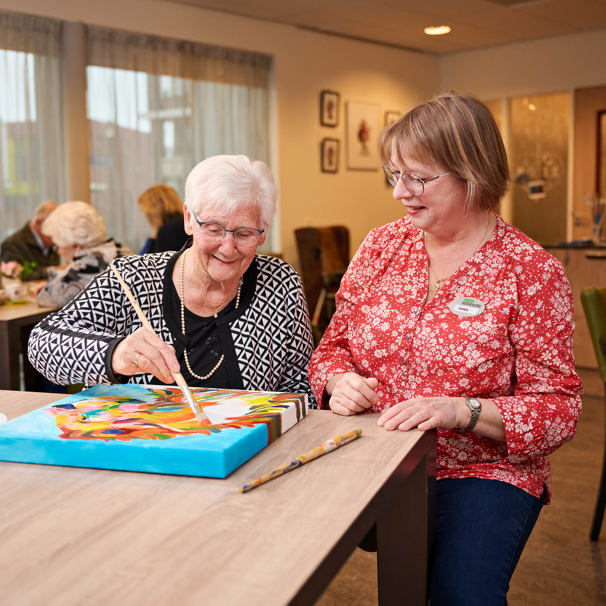 Een oudere mevrouw zit samen met een medewerker van Anna Ouderenzorg aan tafel in een woonzorgcentrum. Ze maakt een schilderij. Op de achtergrond zitten mensen aan andere tafel.  Als je nog thuis woont en graag gezellige activiteiten wil doen of hulp wilt, dan kun je overdag naar de dagbesteding van Anna.