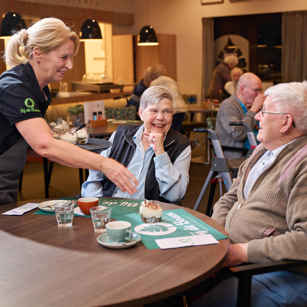 Activiteiten kalender Anna Ouderenzorg Oudere meneer en mevrouw zitten aan een tafel, lachen naar elkaar terwijl een mevrouw toetjes en koffie op tafel zet