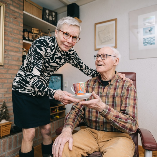 Mantelzorger Nellie geeft kopje koffie aan Frans in de woonkamer. Nellie en Frans van Hoof vertellen over dagbesteding De Groenling bij Anna Ouderenzorg.